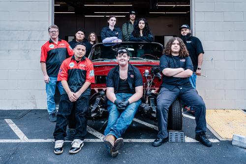 Teen and adult mechanics pose around a red convertible in progress, parked at the entryway of a garage workshop.