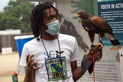 A young pose wearing a face mask and an Earth Conservation Corps T-shirt gestures with one hand as he holds a brown falcon in the other.