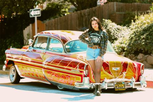 A woman in plaid overshirt and jean shorts, one hand on her hip, poses in front of an intricately painted red and yellow lowrider car parked on the street.