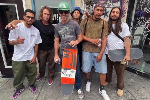 Six people pose outside a storefront and a wall full of graffiti. Two of them hold skateboards.