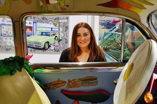 Through the open windows of a class car with flying sandwiches painted in the interior, a woman with long brown hair smiles. The mural behind her also depict a classic car.