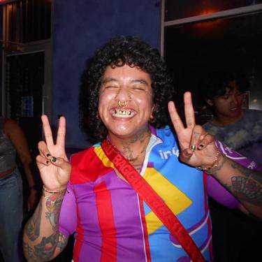 A person with brightly colored shirt, tattooed arms and neck, and gold jewelry smiles broadly while giving two peace signs.