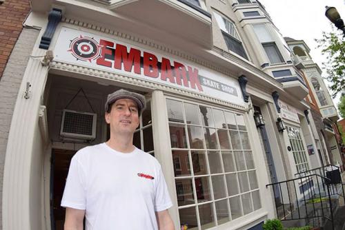 A man poses outside a shope with sign above the door: Embark Skate Shop.