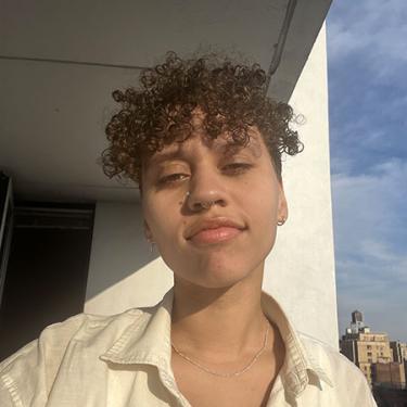 A person with short curly brown hair smiles wryly at the camera.