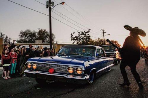 A mariachi plays guitar next to a deep blue lowrider car for a crowd gathered on the other side at sunset.