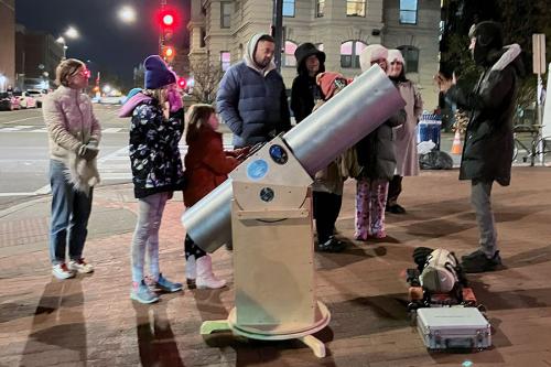 A crowd forms around a large portable telescope on a city sidewalk at night.