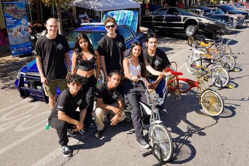 Seven teens pose next to rows of lowrider bicycles and cars parked in a street.