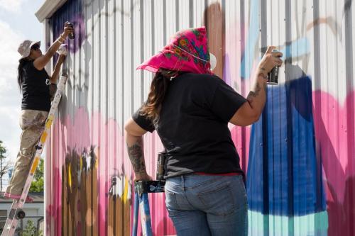 Two people stand on ladders using spray cans to paint an outdoor wall.