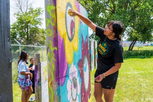 A young woman spraypaints a makeshift wooden wall outside, using bright yellow, purples, and turquoise.