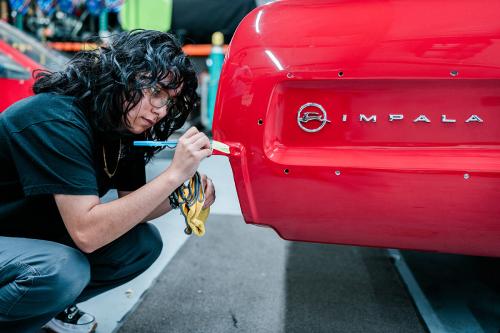 A teenager with wavy dark hair crouches on the ground of a garage, using a ballpoint pen to mark masking tape on the bumper of a bright red Impala.