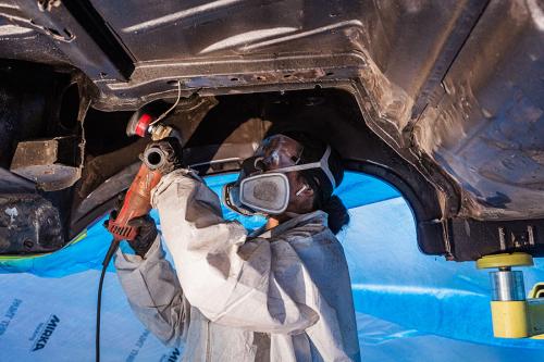 A person in protective gear works with a power tool on the raised undercarriage of a car.