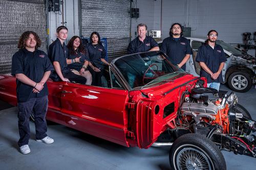 Seven people in matching black work shirts pose in and around a bright red convertible car with the hood removed to show the engine.