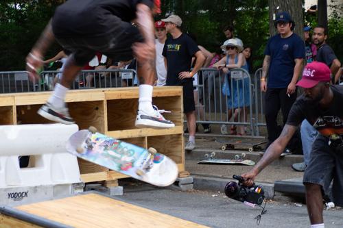 In an outdoor skate course, one person is mid-air in a kickflip while another crotches with a video camera to film them from below.
