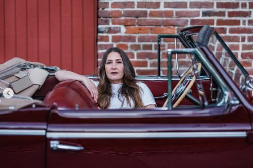 A woman with long dark hair poses sitting across the front seats of a burgundy convertible.
