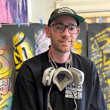 A man smiles wearing a ballcap and respirator around his neck. Behind him are graffiti-style artworks.