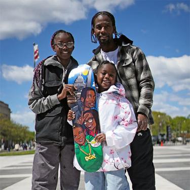 A man and two young girls pose with a skateboard, the bottom of which depicts the three of theme with the Capitol Building and Washington Monument in the background.