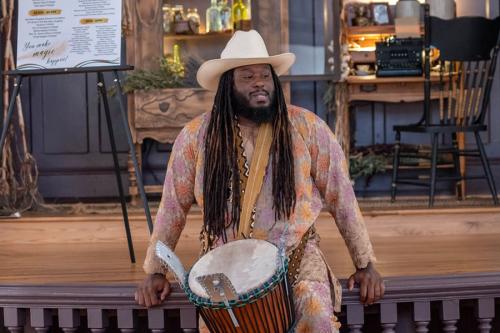 A man with long dark locs and beard, wearing a tan cowboy hat and pale purple and orange outfit, sits at the front of a stage with a hand drum slung over his shoulder.