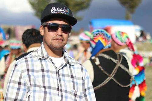 A man in black hat, sunglasses, and light plaid shirt poses in front of people wearing traditional Andean dress, including rainbow-colored woven hats.