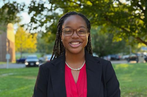 A young woman with long dark braids, glasses, and black blazer over a fucshia blouse smiles.