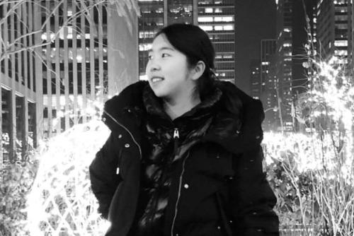 A young woman with dark hair and heavy black jacket smiles, a nighttime cityscape behind her. Black-and-white photo.