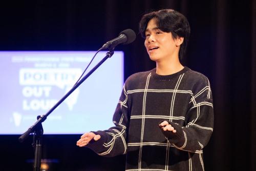 A young man with black hair and black sweater speaks on stage in from of a microphone. A screen behind him reads Poetry Out Loud.