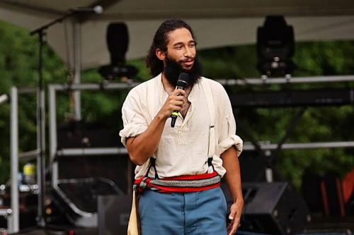 A man with dark hair in a ponytail and bushy beard speaks into a microphone in front of an outdoor stage.