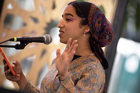 A woman dressed in patterned headscarf and dress stands at a microphone, gesticulating with one hand and holding a smartphone with the other. She wears sparkling purple eyeliner.