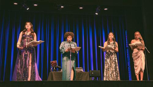 Four young people in long evening gowns stand on stage, each with an open book and a microphone placed in front of them.