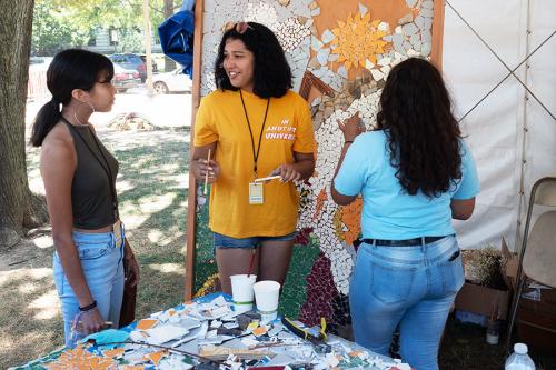 Three people stand in an outdoor festival booth, working on a large tile mosaic. At least two of them wear Festival volunteer badges on lanyards.