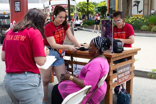 Three young people in matching red Festival shirts interview a woman in pink, seated at a festival booth outdoors.