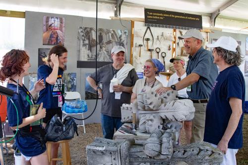 Seven people laughing together while standing under a tent, surrounding a stone carving.
