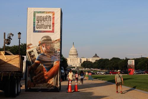 Festival marker with photo of a tradesperson with 'Youth and the Future of Culture' logo towers over National Mall as pedestrians walk by, with Capitol Building in the background.