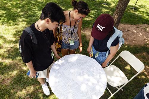 From a high angle, three young people gather around a small circular table that has been decoupaged with pages of poetry.
