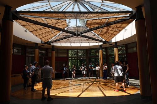 A group of people stand in a circle in a circular room with wooden columns and a circle of sunlight cast through the skylight ceiling.