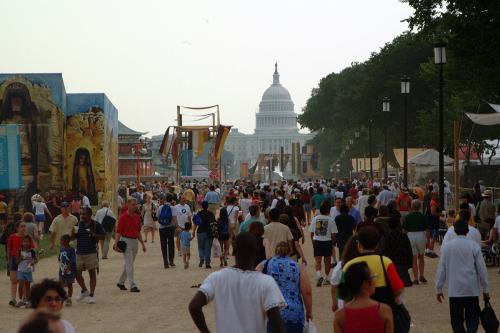Crowds of people on the National Mall facing the U.S. Capitol Building, with festival tents and banners on one side and trees on the other.