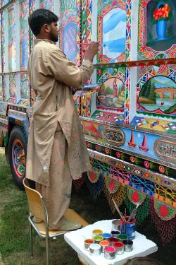 A man with dark skin and tan tunic and pants paints detail in a water landscape image on the side of a decorated truck. A table with open paint cans sits beside him.
