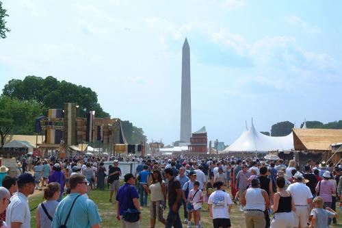 Crowds of people on the National Mall facing the Washington Monument, with festival tents and banners.