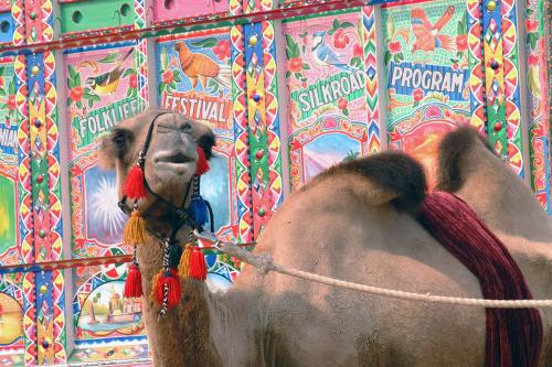 A camel with red tassels on its harness stands in front of a brightly colored mural that reads FOLKLIFE FESTIVAL SILKROAD PROGRAM.