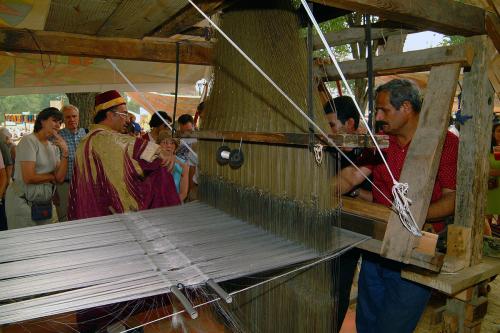 Two men operate a large wooden loom while another explains their process to a crowd of onlookers.