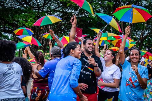 A group of young people dance outside, looking ecstatic, holding up small rainbow-colored umbrellas as confetti rains down.
