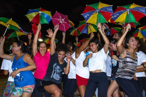 People dancing outside, looking joyous and raising up small rainbow-colored umbrellas. The sky is dark, but they are lit up.