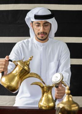 The man pours brown coffee from one brass pot into another smaller one as he smiles slightly.