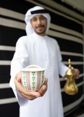 The man holds a brass pot in one hand and holds out a white patterened cup toward the camera in the other. The cup is in focus; he is not.