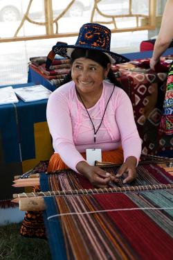 A woman wearing a pink blouse and embroidered hat sits at a loom with colorful fibers.