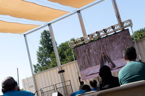 People sit outdoors, watching a video screen.