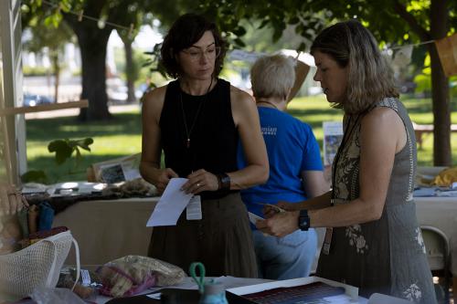 Two women stand at a table under an outdoor tent, each holding pieces of paper or fabric.