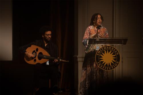 Two people performing on stage: on the left, a man seated playing a stringed instrument; on the right, a woman standing at a podium speaking.