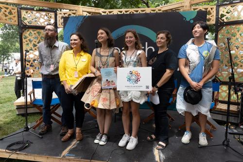 Six people stand on a small, outdoor stage, holding up a sign that reads “Global Co Lab Network” with a rainbow-colored logo. Three of the individuals are dressed in upcycled trash outfits made of paper grocery bags, N-95 masks, and silver packaging mater