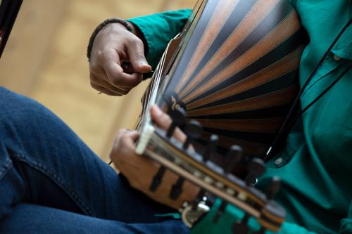 Hands pluck the strings of a striped oud. The musician is seated and wearing a blue-green shirt.