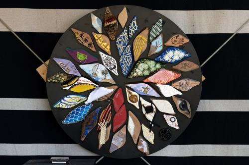 On a black circular board, several eye-shaped pieces of painted wood, arranged as if bursting outward. Each is designed differently, including blue with gold calligraphy, a realistic eye in pastels, coffee beans glued to the surface, a white fishing net o